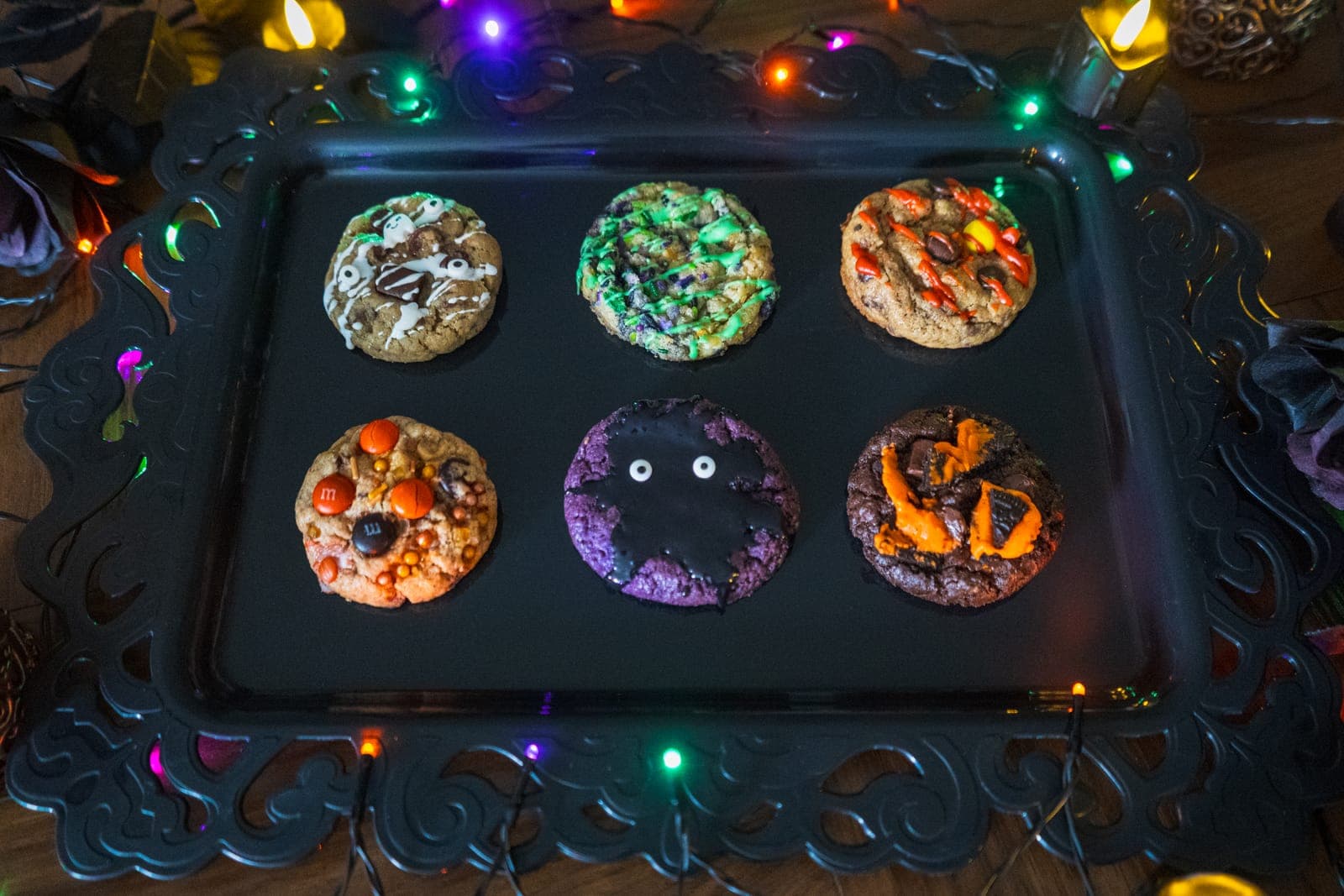 Six Halloween cookies on a black tray: Spooky Chocolate Chip, Slimy Sugar, Bloody Reese’s, Fall M&M, Ube Monster and Oreo Brownie