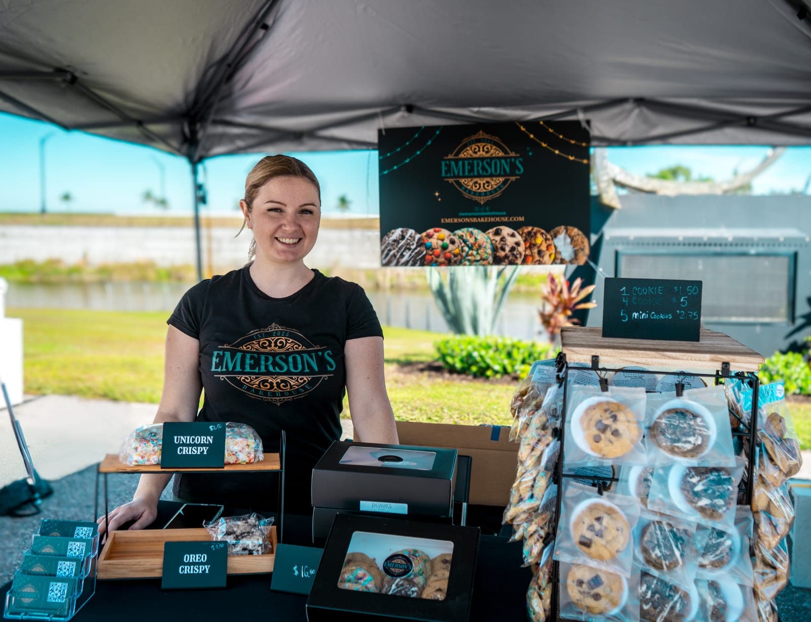 Sarah smiling behind the Emerson's Bakehouse market table with cookie boxes, crispies and a cookie rack